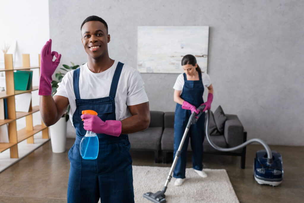 cleaner showing okay and holding detergent near colleague using vacuum cleaner